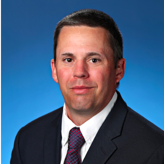 professional headshot of white male in black suit jacket and red/blue tie against a blue background